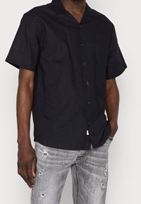 Kurzarm, dunkelblaues Hemd mit Knöpfen aus leichtem Stoff. Mit einem patrizierten Kragen und subtiler Glanzoptik. Kombiniert mit verwitterten grauen Jeans.