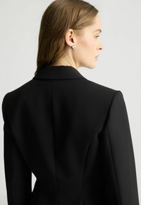 Blazer nero su misura con spalle strutturate, tessuto liscio e chiusura a un bottone, visto da dietro con un accenno di gioielli per orecchie.