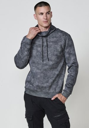 Sudadera gris con un patrón texturizado, cuello alto y cordones. Puños y dobladillo acanalados, combinada con pantalones cargo oscuros.
