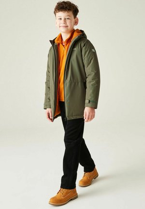 Chłopiec idący w oliwkowozielonej kurtce z kapturem, pomarańczowej polarowej bluzie, czarnych spodniach i beżowych butach, na tle jasnego, jednolitego tła.