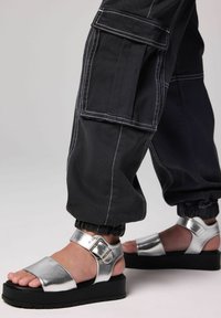 Hopeiset metalliset sandaalit sisältävät leveän vyön, säädettävän soljen ja paksun mustan alustapohjan. Yhdistetty mustiin cargo-housuihin.