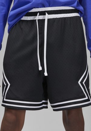 Schwarze Basketballshorts aus Netzstoff mit weißem Kordelzug und weißen Streifendetails, getragen mit einem blauen Hemd am unteren Torso und den Beinen einer Person.