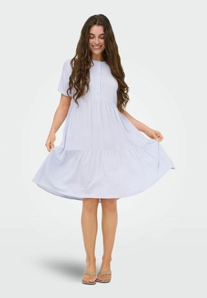 Femme aux longs cheveux bouclés portant une robe bleu clair à manches courtes boutonnée, tenant son ourlet et souriant, debout pieds nus.
