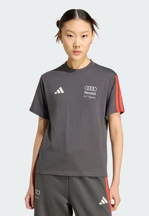 Giovane donna che indossa una maglietta grigia Adidas Revolut F1 Team e pantaloni coordinati con strisce rosse, in piedi contro uno sfondo chiaro.