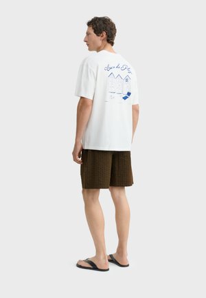 Mann trägt weißes T-Shirt mit blauem Strandhütten-Design und Text, braune, strukturierte Shorts und schwarze Flip-Flops, steht vor weißem Hintergrund.