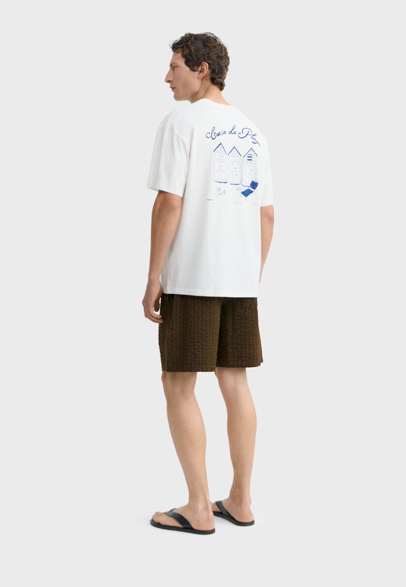 Homme portant un t-shirt blanc avec un motif de cabine de plage bleue et du texte, un short marron texturé, et des tongs noires, debout devant un fond blanc.