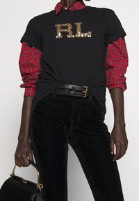 Camiseta negra con el logo "RL" en lentejuelas doradas, superpuesta sobre una camisa de cuadros en rojo y negro, combinada con pantalones de terciopelo negros y un bolso negro pequeño.