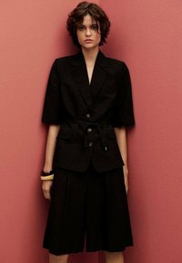 Black linen jacket with short sleeves, belted waist, large buttons, and front pockets, paired with tailored black shorts. Background is pink.