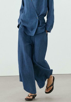 Persona che indossa pantaloni blu larghi e blazer abbinato, cammina con sandali neri aperti con tacco su un pavimento liscio.