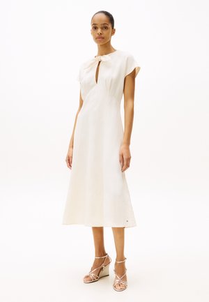 Robe crème avec un corsage ajusté, encolure à trou de serrure, manches courtes et jupe évasée en A ; accentuée par un détail en nœud.