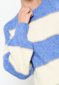 Kootud sviiter, millel on sinised ja kreemja triibud, tekstuuriga viimistlus ning dekoratiivne kuldne õmblus šeffide ääres.