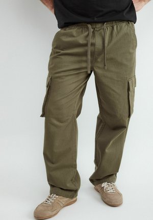 Mężczyzna noszący oliwkowo-zielone spodnie bojówki z bocznymi kieszeniami i beżowe sneakersy, stojący na jednolitym tle.