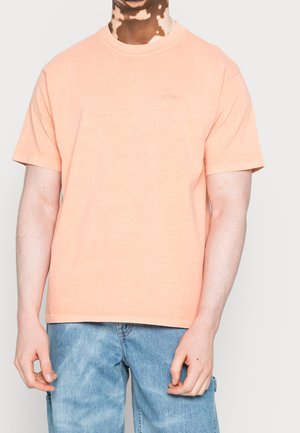 Homme atteint de vitiligo portant une chemise à manches courtes couleur pêche clair et un jean bleu en denim sur un fond uni.