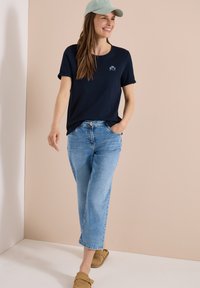Mujer sonriente caminando con camiseta negra, vaqueros cortos azul claro, zapatos beige sin cordones y una gorra de béisbol verde claro sobre fondo beige.