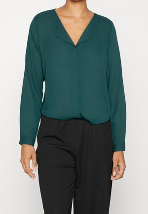 Persoon draagt een donkergroene blouse met lange mouwen en een V-hals, en zwarte hoge taille broeken, staand tegen een effen achtergrond.