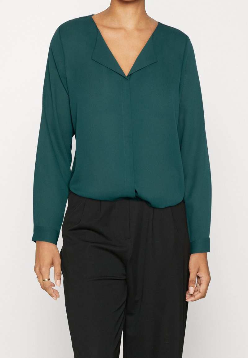 Personne portant un chemisier vert foncé à manches longues avec un col en V et un pantalon noir taille haute, debout devant un fond uni.