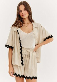 Lněný outfit obsahující béžovou košili s krátkým rukávem a černým vlnkovaným lemem a odpovídající šortky s elastickým pasem. Svetřík pod tím.