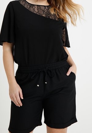 Kobieta ubrana w czarny strój z koronkowymi detalami na górze i szorty z troczkiem, jedna ręka w kieszeni na jednolitym jasnym tle.