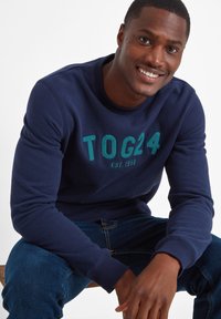 Navy sweatshirt with crew neck, featuring textured green "TOG24" logo. Paired with blue denim jeans. Smooth fabric and ribbed cuffs.