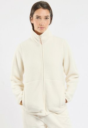 Femme portant une veste en polaire de couleur crème avec un col montant et une fermeture éclair à l'avant, les mains dans les poches, regardant devant elle.