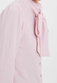 Blusa texture rosa chiaro con collo alto e dettagli a fiocco annodato, caratterizzata da maniche a sbuffo e chiusura a bottoni lungo la parte anteriore.