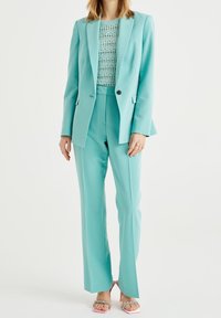 En teal kostym består av en skräddarsydd kavaj och passande byxor. Under den bärs en stickad topp som tillför struktur. Outfiten kombineras med rosa klackar.