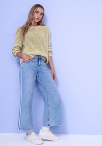 Femme portant un pull tricoté jaune-vert, un jean large bleu clair avec des bandes latérales, des chaussettes blanches et des baskets blanches, debout contre un mur violet.