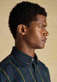 Mladý muž s krátkymi kučeravými vlasmi, oblečený v zapnutej tmavej košeli s vertikálnymi žltými, zelenými a červenými pruhmi, stojí otočený doprava.