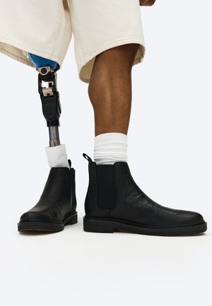 Personne portant un short beige et des chaussettes blanches, avec une jambe naturelle et une jambe prothétique, toutes deux chaussées de bottines Chelsea noires.
