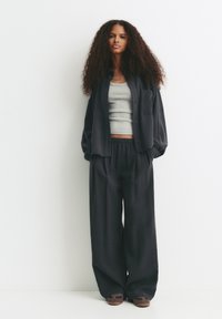 Woman with long curly hair wearing a loose dark shirt and wide black pants over a grey tank top, standing against a white background.