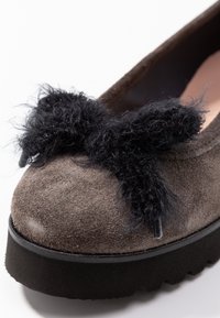 Zapato de ante gris con una gruesa suela de goma negra. La puntera presenta un prominente lazo negro y esponjoso, que añade textura al diseño.