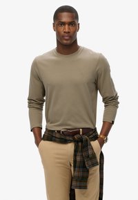 Zielona oliwkowa koszula z długim rękawem i okrągłym dekoltem, z małym logo. Beżowe spodnie, brązowy pasek i koszula w kratę związana w pasie.