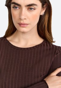 Maglione marrone a coste con maniche lunghe e scollo rotondo. Tessuto liscio, design aderente e leggero lucido. Il modello ha i capelli ben pettinati.