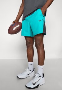 Shorts de sport turquoise et gris avec un logo Nike, associés à des chaussures Nike blanches et des chaussettes texturées, tenant un ballon de football brun.