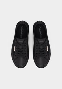 Ζευγάρι μαύρα αθλητικά παπούτσια Calvin Klein με πλέγμα, κορδόνια, φιξάκια με το σήμα στις πλευρές και ορατό λογότυπο στις σόλες και τη γλώσσα, εμφανιζόμενα από πάνω.