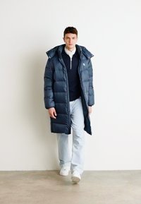 Marineblaue Steppjacke mit langem Design, Kapuze und gestepptem Muster; getragen über einem dunklen Pullover und hellblauen Jeans; weiße Sneakers.