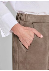 Bruine suède broeken met een hand in de zijzak. De tailleband is elastisch en de stof heeft een zachte, gestructureerde afwerking.
