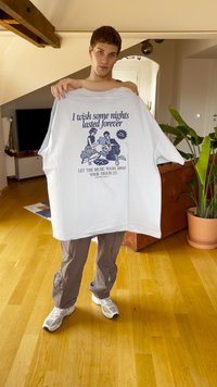 Balts oversized T-kreklis ar grafisku apdruku, kurā attēlotas divas figūras un teksts. Pelēki kravas bikses un baltas sporta kedas ar melniem akcentiem. Iekštelpu vide.