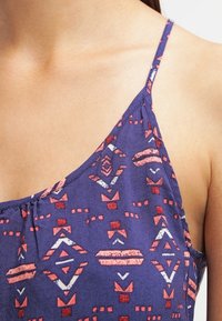 Haut en tissu violet avec un motif géométrique comportant des accents rouges et blancs. Bretelles spaghetti ajustables et design froncé au niveau du décolleté.