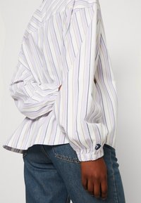 Camicia a maniche lunghe a righe in bianco, lilla e oro, con una vestibilità rilassata, polsini arricciati e orlo curvo. Indossata con jeans blu scuro.