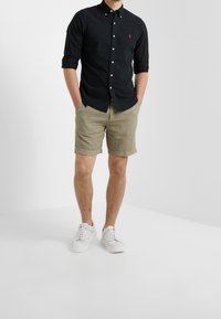 Černá košile na knoflíky, khaki šortky a bílé tenisky. Košile má malé logo; šortky jsou na lemu ohrnuté, vyrobené z lehkého materiálu.