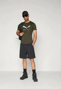 Camiseta de manga curta verde oliva com logo, calções pretos e botas pretas. O modelo carrega uma mochila preta e usa óculos de sol e um boné.