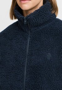 Veste en polaire bleu marine foncé avec col haut, fermeture éclair à l'avant et surface texturée. Présente un logo discret sur le côté droit inférieur.