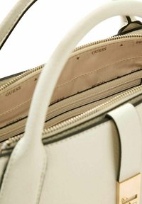 Sac à main en cuir blanc avec des finitions en métal doré. L'intérieur présente une doublure beige avec le nom de la marque imprimé. Compartiment zippé et plusieurs poches visibles.