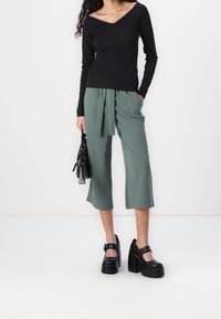 Person, der ein schwarzes Langarmoberteil und eine grüne, verkürzte Hose mit Gürtel trägt, eine schwarze Handtasche hält und schwarze Plateauschuhe trägt.
