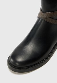 Bottine noire en cuir lisse avec bout arrondi et détail de sangle marron texturée sur la partie supérieure.