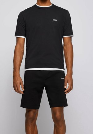 Homme portant un t-shirt noir à manches courtes et un short noir, tous deux avec des bordures blanches et un petit logo "BOSS" sur la poitrine et la cuisse.