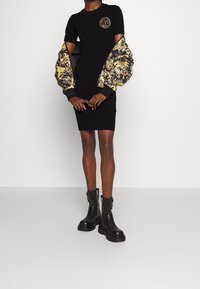 Svart bodycon-klänning med rund halsringning, pryds av överdimensionerade mönstrade ärmar i guld och svart. Kombineras med kraftiga svarta ankelskor.