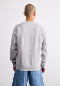Felpa grigia con collo rotondo e polsini a coste, caratterizzata da una vestibilità comoda. Indossata con pantaloni di jeans chiari, che mostrano una trama liscia.