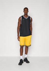 Athletisk mand stående iført sort ærmeløs sportsjersey, gule basketballshorts, hvide sokker og sorte sneakers foran en ensfarvet baggrund.